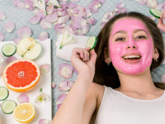 Spa -skin- care- cosmetology- concept- Pretty -smiling- woman -with -pink -clay -facial- mask -and -cucumber- slices -in- her -hands- lying- on -the-bed -flower-- petals -and -fresh -fruits -around -her
